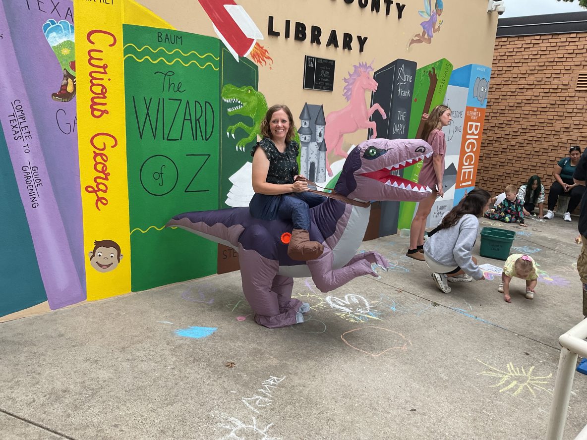 librarian in a dinosaur inflatable costume stands in front of library mural while children chalk on the porch