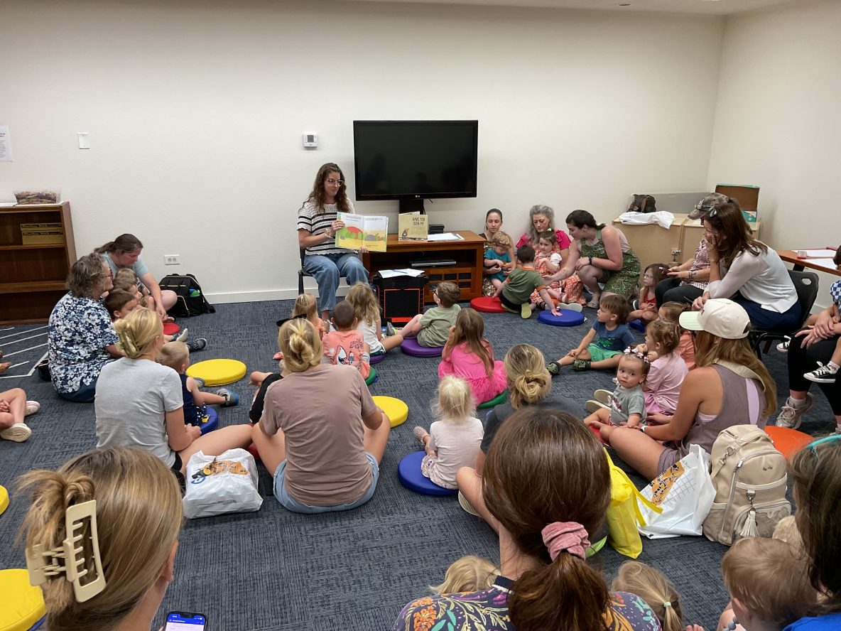librarian reads a picture book about dinosaurs to a large group of children and caregivers