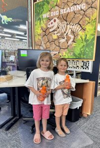 two girls stand in front of library computers holding their summer reading prizes.