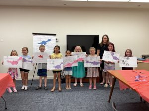 Students hold their dinosaur artwork in the library's meeting room.