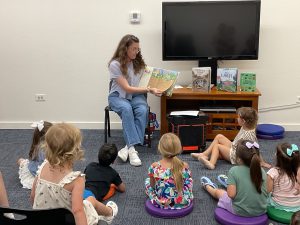 Seated librarian reads a picture book about worms to a group of children sitting on colorful cushions.