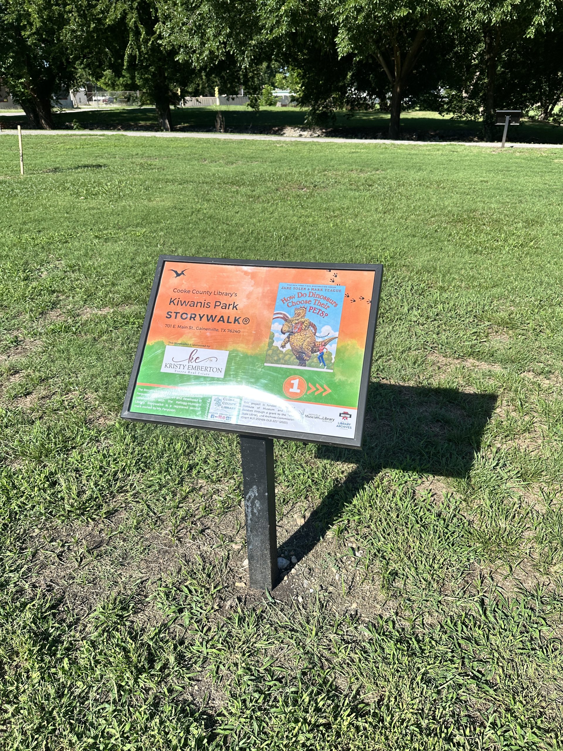 Image of a display case in a grassy park holding the title page of Jane Yolen's picture book "How Do Dinosaurs Choose Their Pets?"