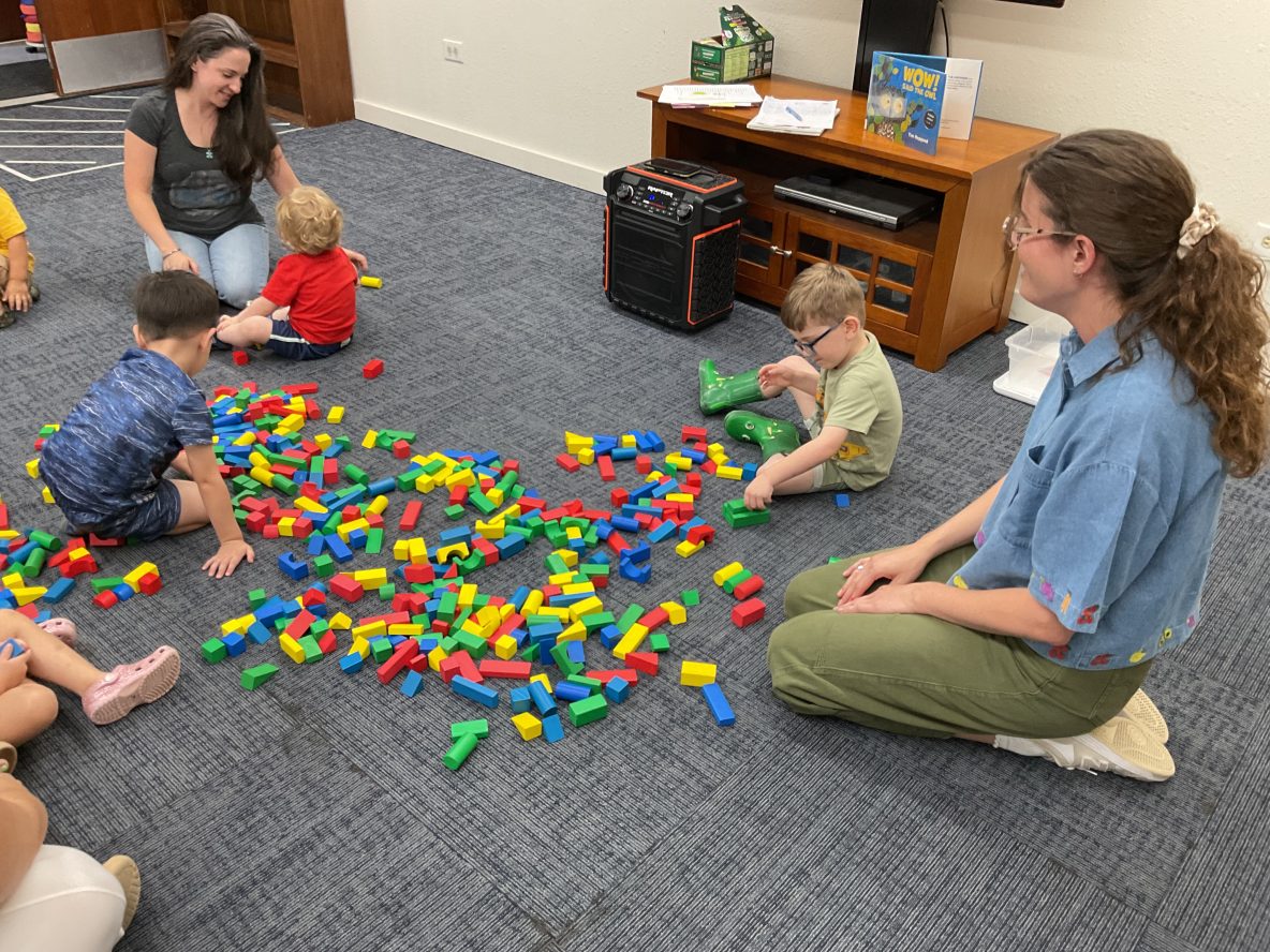 library meeting room with seated adults and children on the floor playing with big duplo blocks
