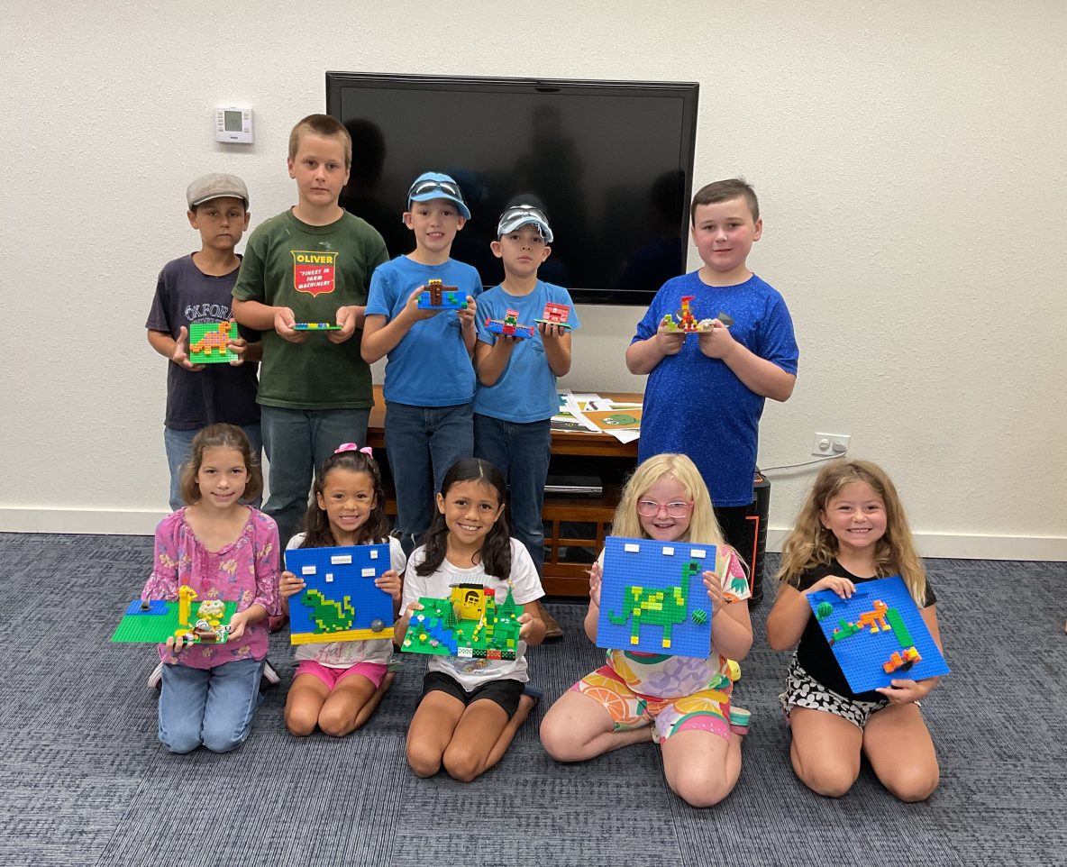 A group of children pose in two rows holding up Lego creations. Most of the children are smiling big and created dinosaurs.