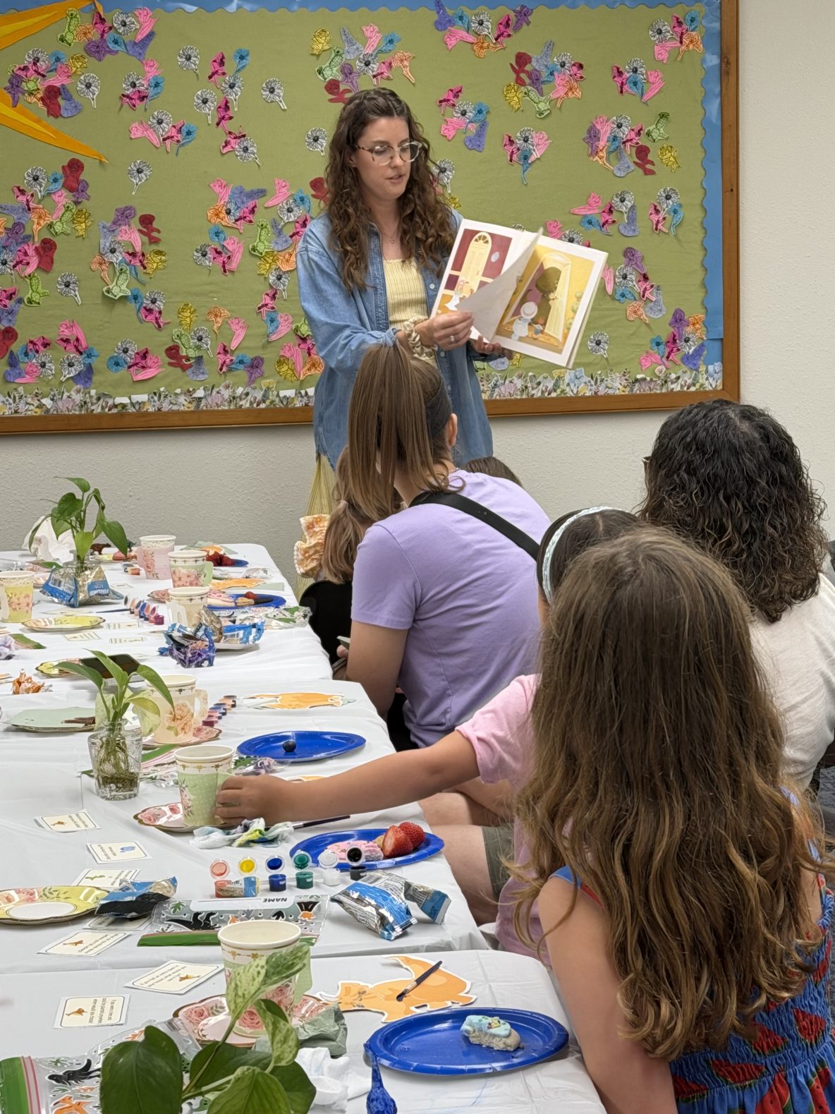 librarian reads "Tea Rex" by Molly Idle to a group of children and caregivers who are seated enjoying tea and treats
