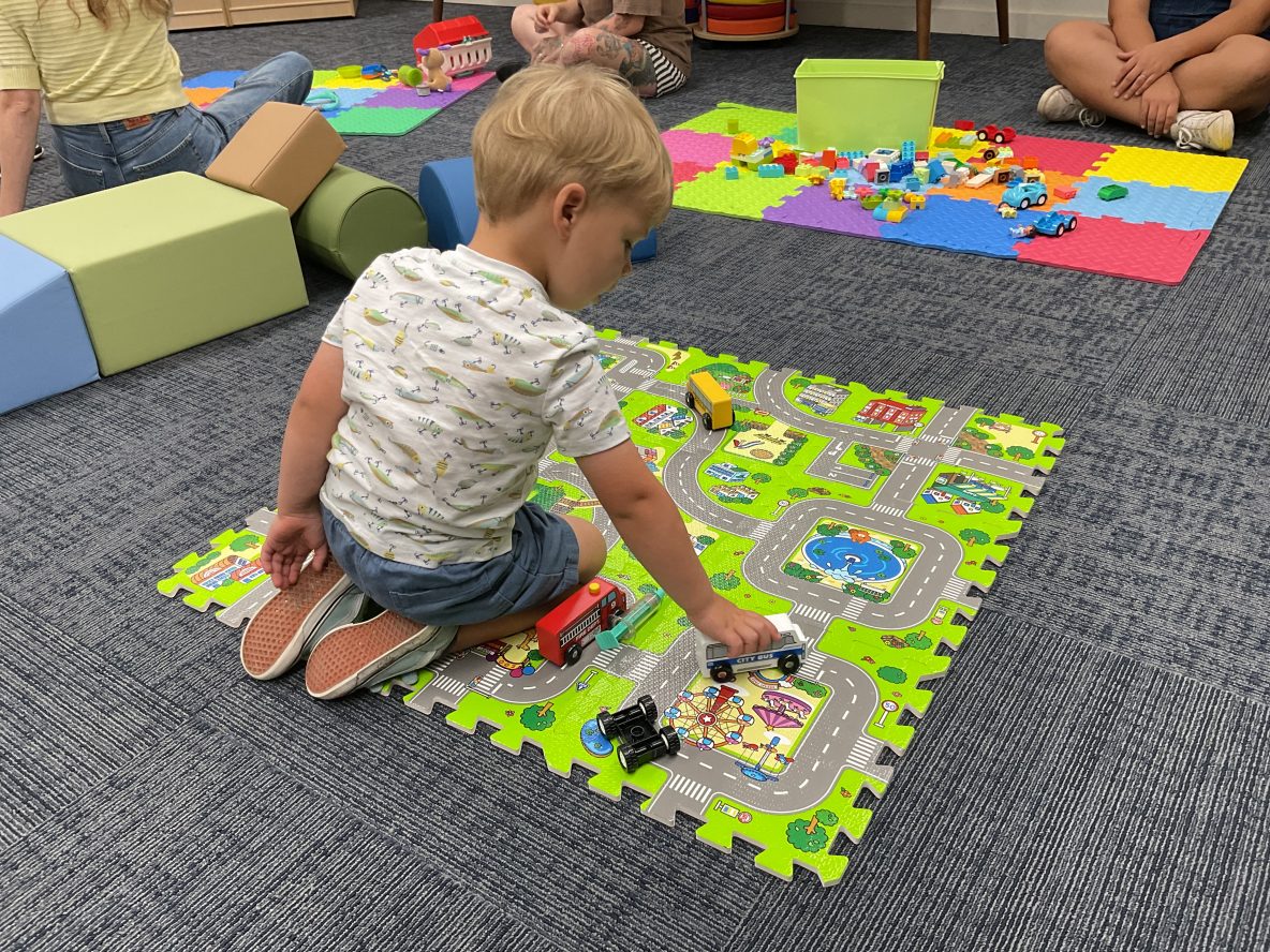 a little toddler is sitting on a mat playing with wooden vehicles. adults are seated around the room behind him and there are various toys for children