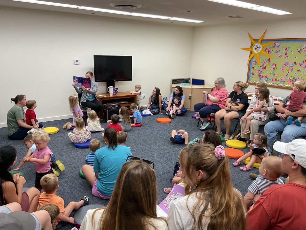 photo of the interior of a library with a librarian reading a picture book to a large group of seated children and adults