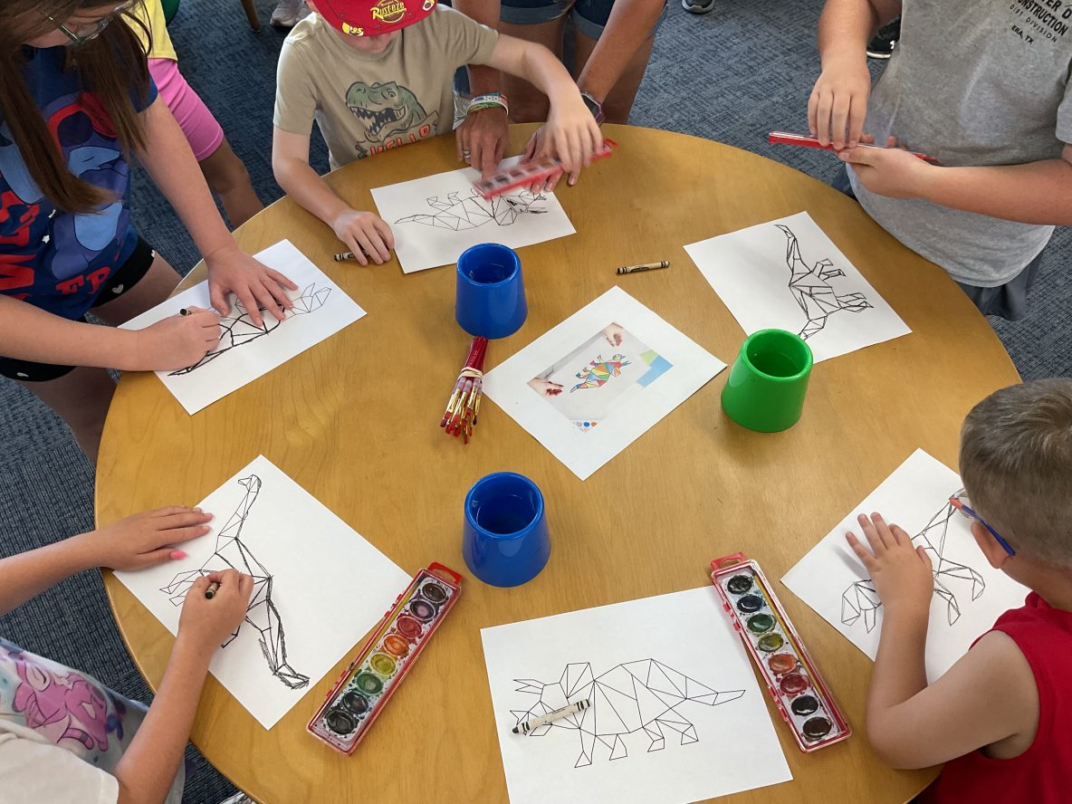 a group of children stand around a table painting dinosaurs with watercolors
