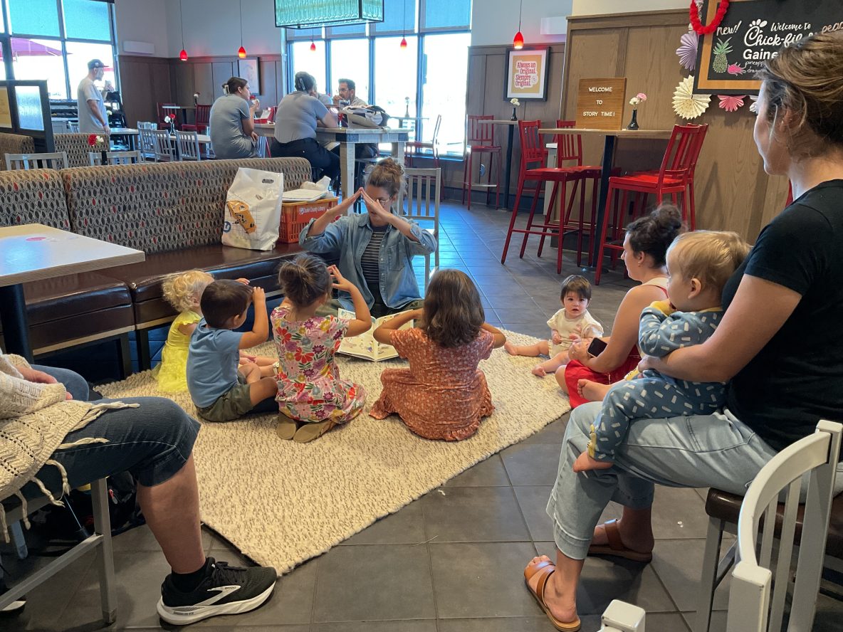 librarian sits on a rug with several children at chickfila. all are making a tent with their hands.