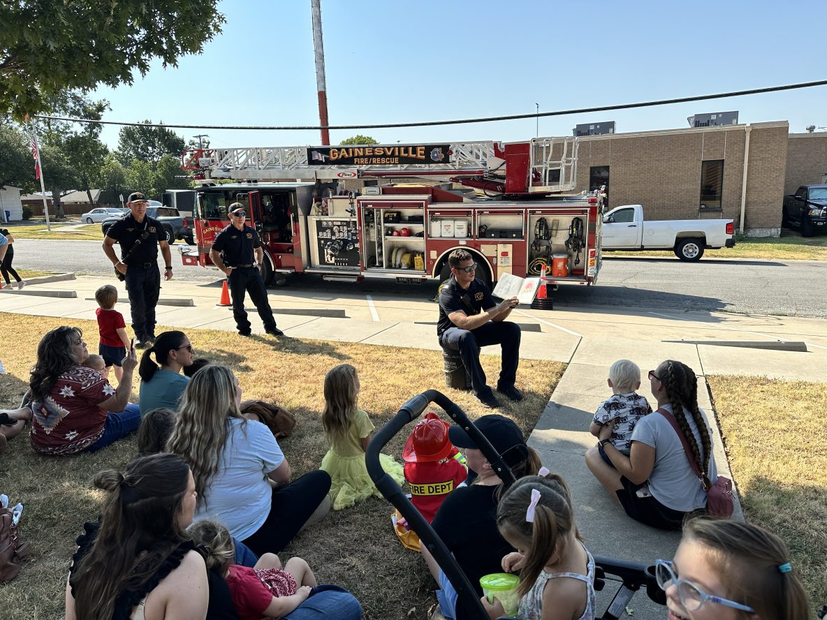 firefighter reads a curious george book about firefighters to a group of children in front of a firetruck