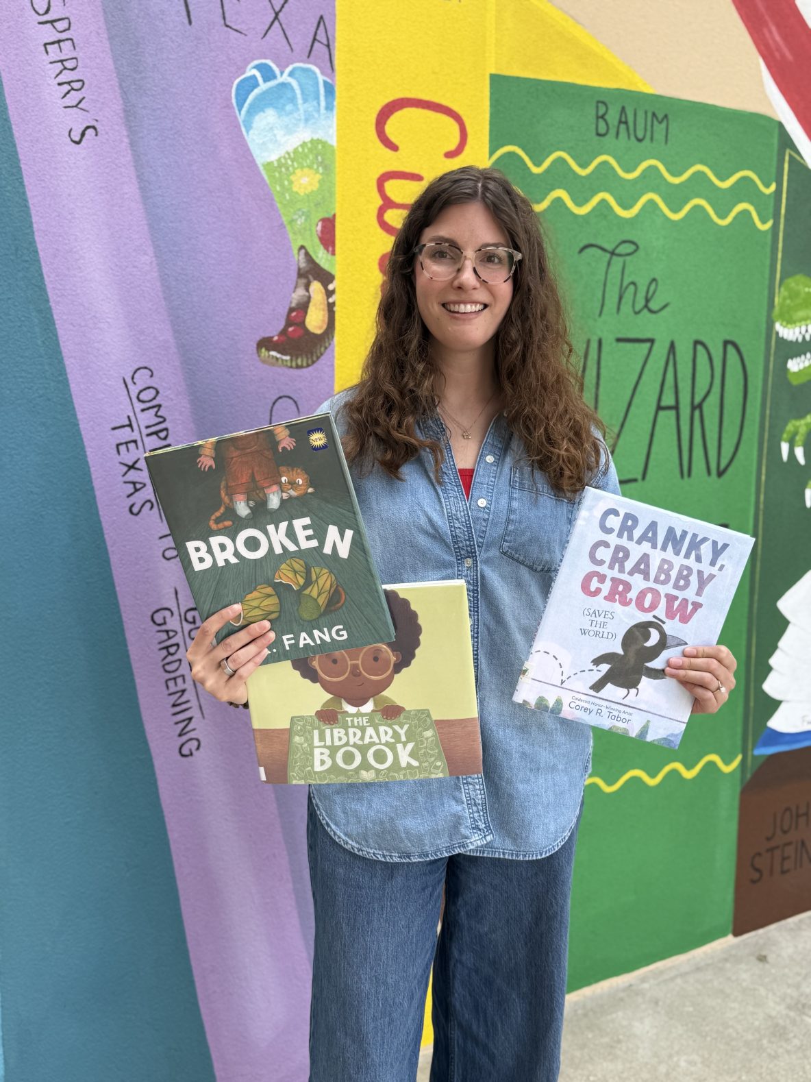 librarian holds copies of "The Library Book," "Cranky Crabby Crow Saves the World," and "Broken" in front of library mural